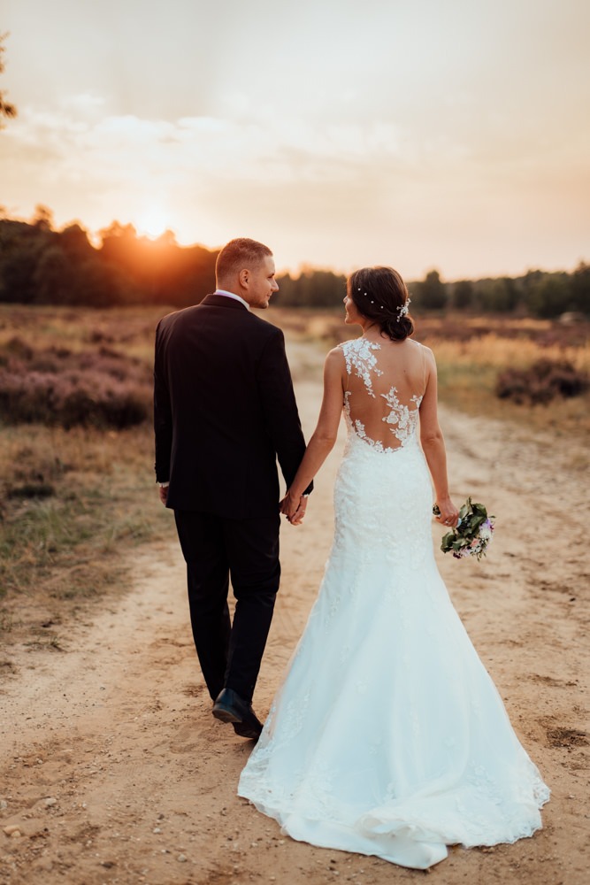 Brautpaar geht auf sandigem Boden Händchen haltend durch eine verträumte Heidelandschaft. Die Sonne geht gerade hinter den Bäumen unter.
