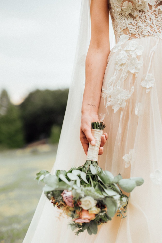 Detailshot von einem wunderschön verzierten Hochzeitskleid und einem Brautstrauß in Pastelltönen. Tätowierte Braut mit zierlichem Schriftzug am Handgelenk.