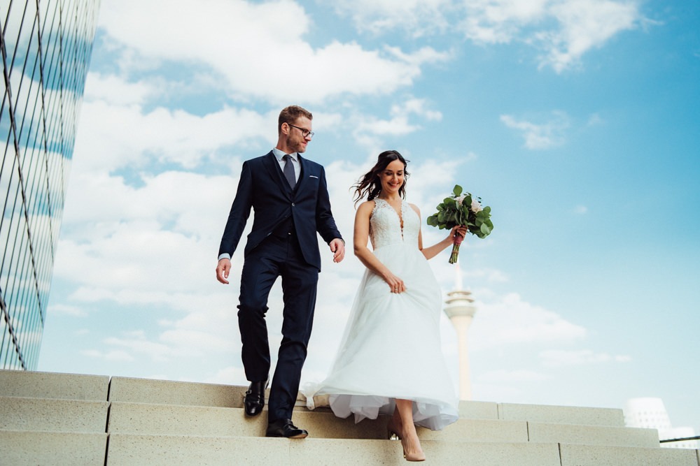Brautpaar am Düsseldorfer Hafen mit Strahlend blauem Himmel. Beide schreiten eine Treppe herunter, Wind weht durch ihre Haare.